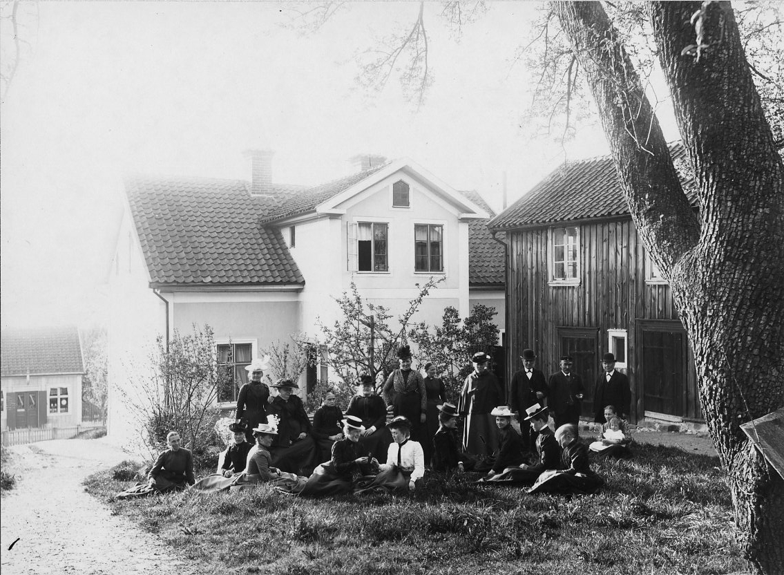 Calla Helena Sundbeck, En större grupp med män, kvinnor och enstaka barn, står och sitter på gräsplan på gården till Brahegatan 40. En putsad byggnad med ett öppet fönster och öppen dörr. Till höger en uthusbyggnad i två våningar med locklistpanel, möjligen rödfärgad.