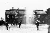 Brandkåren har övning på Stora torget. På torget står ett barn och pratar med en uniformsklädd man, även en ensam man i stråhatt går över torget. I bildens mitt står brandchef Alfred Hedén.