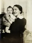 Judit Johansson (1893 - 1973) sitter i en fåtölj och håller sonen Thure Johansson Bjerrhede (1918 - 2004) cirka 1918-19. De bodde i Vommedal Östergård. Motivet är taget i Judits svärföräldrars hem Alegårdsslätt, Fässberg.