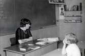 Lärare Ruth Singmyr sittandes vid en kateder i Kålleredskolan/Brattåsskolan cirka 1965. Bakom henne ses 