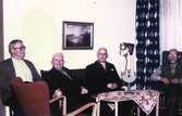 Fyra män sitter vid ett bord i dagrummet på första våningen i Brattåshemmet innan 1960. Från vänster ses skräddaren Johan Blomkvist (1887 - 1969), okänd man, Oskar Andersson (1877 - 1960, från Kungsbacka) samt drängen Robert Pettersson (1886 - 1968).
