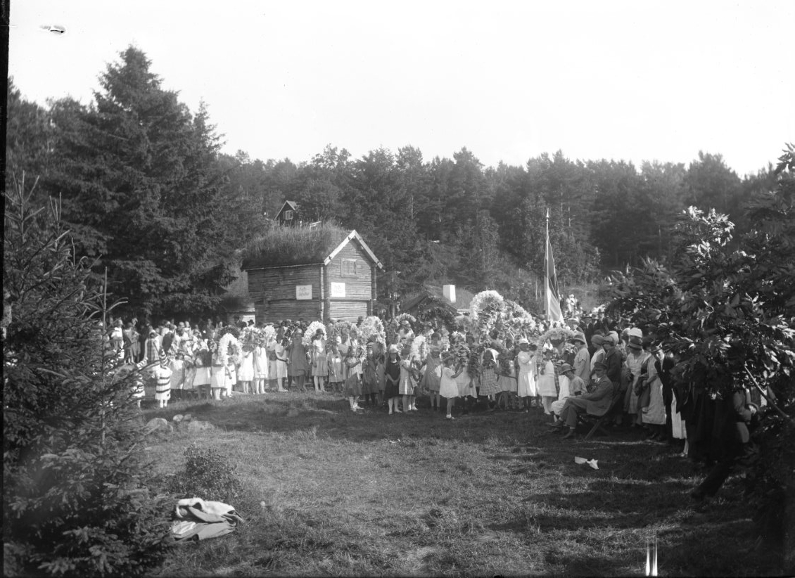 Calla Helena Sundbeck, Folksamling, barn och vuxna vid Friluftsmuseet på Grännaberget, det är Hembygdsfest. Barnen håller blomsterbågar i händerna. De står framför pappersmakarbostället vars loftbod har en skylt: "Kaffebiljetter"