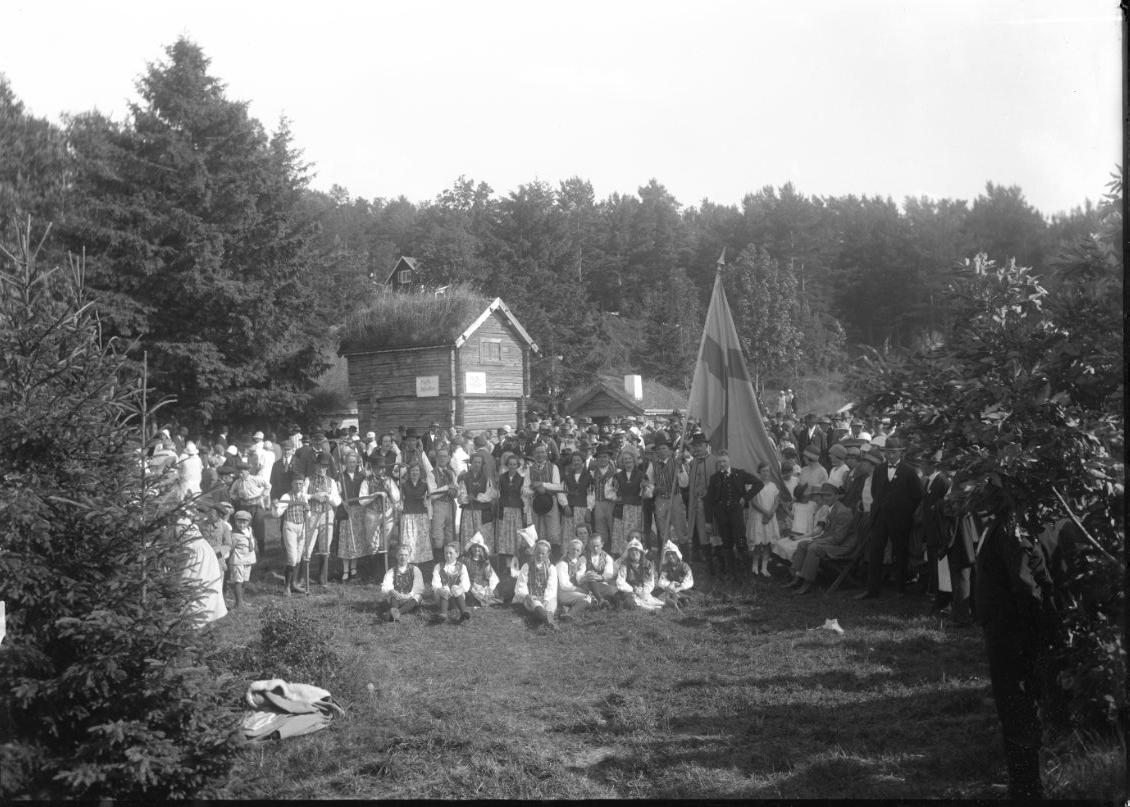 Calla Helena Sundbeck, Folksamling, barn och vuxna vid Friluftsmuseet på Grännaberget, det är Hembygdsfest. Barnen håller blomsterbågar i händerna. De står framför pappersmakarbostället vars loftbod har en skylt: "Kaffebiljetter". I ett främre led står och sitter vuxna och unga folkdansare. Det bakre ledet har liar i hän…