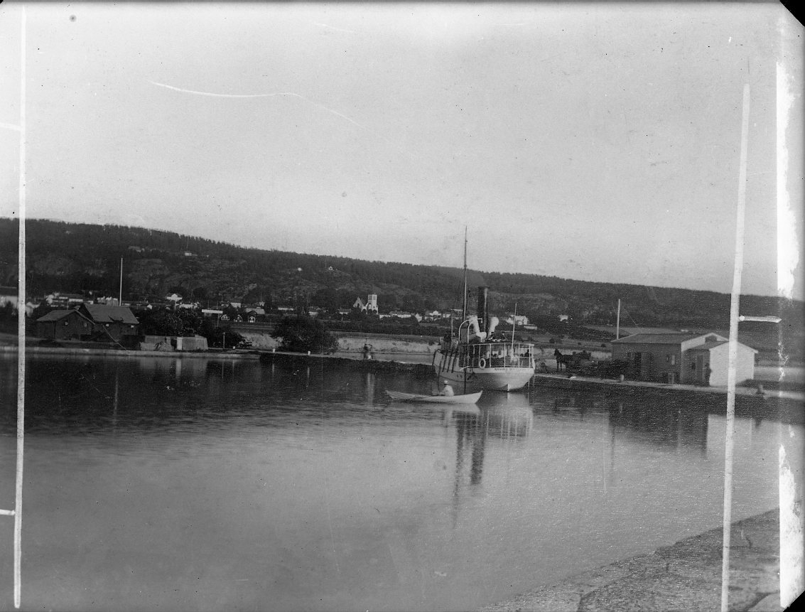 Calla Helena Sundbeck, En ångbåt ligger vid kaj i Gränna hamn. En roddbåt passerar. Ångbåten är identifierad som Södra Wettern som gick regelbunden trafik mellan Gränna och Visingsö.