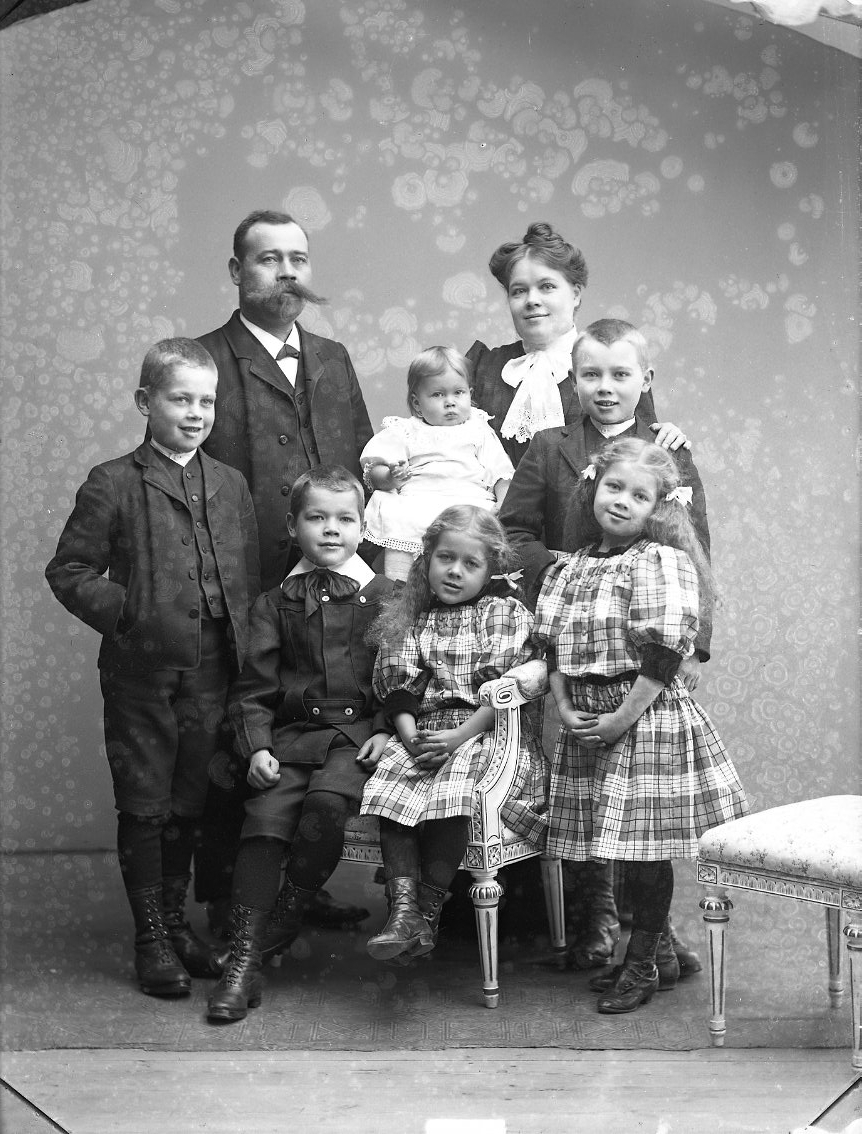 Calla Helena Sundbeck, Gruppbild med familjen Johansson. Sex barn - tre flickor och tre pojkar - står och sitter framför herr och fru Johansson. De äldre flickorna har storrutiga klänningar och rosetter i håret. De ler.