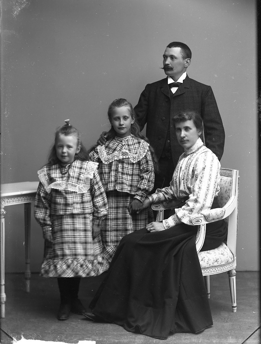 Calla Helena Sundbeck, Gruppbild med familjen Franzén i Gränna. Två flickor i storrutiga klänningar med volang står intill deras mamma som sitter i en stol av gustaviansk stil. Flickan närmast mamman (den äldsta) håller hennes hand. Mannen står bakom dem. Hans högra hand vilar på den äldre flickans högra axel. Han tittar…