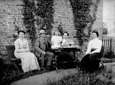 EN MAN, TRE KVINNOR OCH ETT BARN DRICKER KAFFE I TRÄDGÅRDEN.