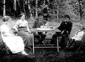 ETT BARN, TVÅ KVINNOR OCH TVÅ MÄN DRICKER KAFFE VID TRÄDGÅRDSBORDET.