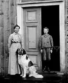 KVINNA OCH POJKE STÅR I DÖRREN TILL ETT BONINGSHUS, FRAMFÖR DEM SITTER EN HUND.