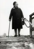 80-åriga Elvira Jägnert (född Johansson i Heljered 1906 - död 1994) promenerar med käpp från Brattåsgården (eller skolan) år 1986. Hon håller i trappräcket på väg ner till tunneln som går under Streteredsvägen mot Våmmedal.