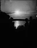 Siluett av kvinnor och män i motljus vid vattendrag, Östhammar, Uppland
