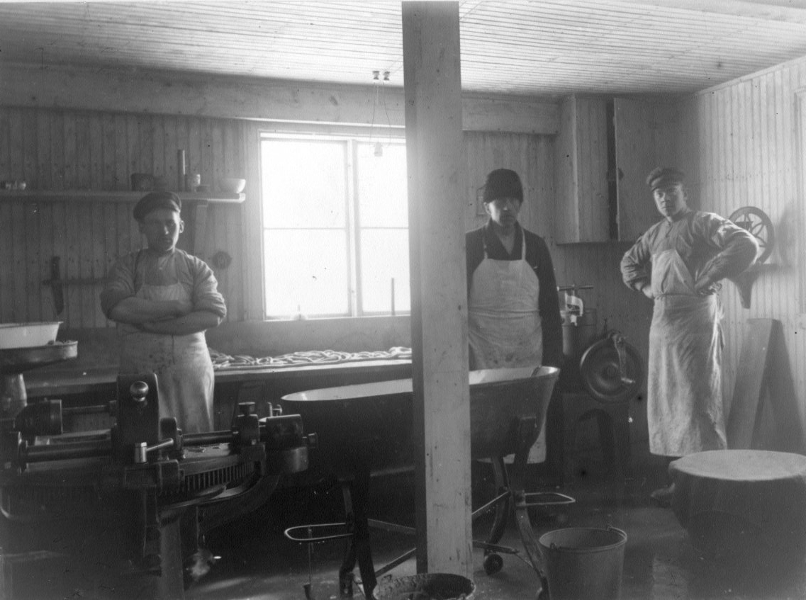 Calla Helena Sundbeck, Interiör från Frans Augustssons Charkuteriaffär i Gränna. Tre män i ljus förkläden och keps/mössa på huvudet står framför ett fönster med starkt solljus. De är från vänster Hugo Andersson, Frans Augustsson och Bengt Skoglund. På en bänk bakom dem ligger korv i långa rader och runtom dem står olika …