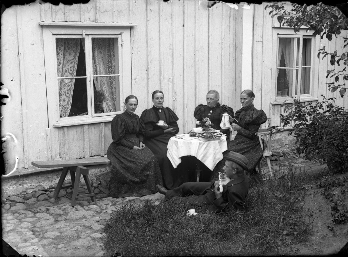 Unknown artist, Coffee drinking in a garden. Four women sit around a table with a coffee cup in their hands, a man with a hat is on the lawn next door with a cat in the fist. A coffee cup stands in the grass next to him. The women on the left and right are possibly the sisters Grönberg.