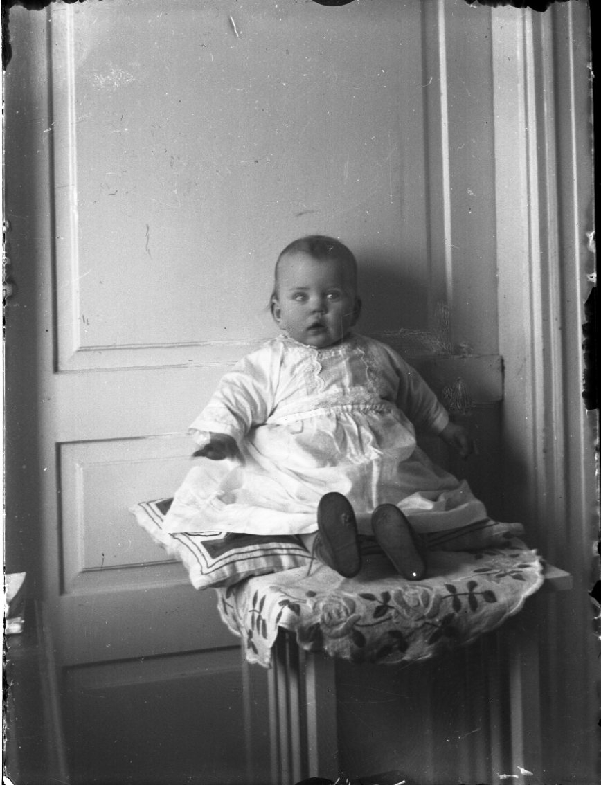 Unknown artist, Photograph of unknown infant sitting on a pillow on a small table leaning against a door.