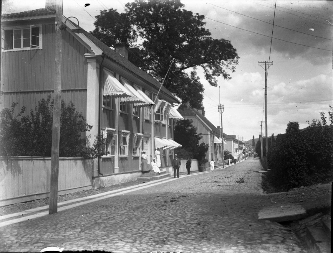 Unknown artist, Prästgårdsbostället vid Brahegatan i Gränna. Utfällda markiser på bägge våningsplanen. Tre kvinnor i ljusa klänningar står respektive sitter på trappan. En man står nedanför, flera personer i rörelse på de två rader med borgmästarsten som utgör trottoaren. Gatan är belagd med kullersten.