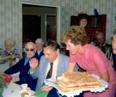 Kokerskan Ingela Sjöberg serverar tårta under en anhörigträff på Brattåshemmet 1979. Från vänster ses Knut Hagberg (i mörka glasögon), Alfred Mattsson (skymtar i bakgrundens mitt) samt Thure Bjerrhede (Knuts släkting från Vommedal). Relaterat motiv: A1892.