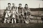 Rast i jordbrukandet på några stockar. Kvinnorna håller i var sitt barn och männen som heter Per-Olof och Bertil Sjögren håller i spadar och grep. Kvinnan till vänster heter Signhild Andersson och flickan till höger är Elin Sjögren.