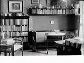 Interiör med bokhyllor och soffgrupp på ena bildedn. Bild 2 (GB2_2728) visar ett rum med mörka väggar fyllda av småtavlor och ljust målade trämöbler i stram jugendstil. Blombuketter hänger i taklampan och står i vaser på bordet, som täcks av en julduk. Ovanför den lilla soffan sitter en långsträckt bonad med julmotiv. Läroverksadjunkt Essén beställde bilderna, som troligen är tagna hos honom, se även bildnr GB2_2729.