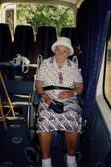 Boende från Brattåsgårdens äldreboende är på bussutflykt till Hembygdsgården Långåker, 1990-tal. Hulda Olsson, iklädd vit hatt och svart/röd-mönstrad vit klänning (1993 - 2000). Hon sitter i en rullstol som står i bussen. Hulda var född i Vommedal Östergård 