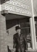 En okänd äldre man, iklädd skinnjacka, käpp och hatt, står framför servicehuset Brattåsgården på Streteredsvägen 5 år 1986.