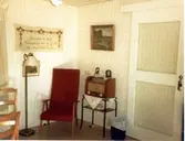 Brattåsgården vid invigningen år 1986. Utställning av föremål från gamla Brattåshemmet, gjord av Kållereds Hembygdsgille. Delar av föremålen finns nu på Mölndals stadsmuseum. Här ses en röd fåtölj, radio på ett litet bord, väggbonad och tavla.