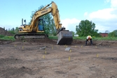 En arkeolog arbetar vid grävmaskinen och i förgrunden finns anläggningar markerade med gula pinnar. Foto taget vid en arkeologisk undersökning i Gränna 2009.