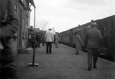 Ett tåg står på spåret och en stins och några resenärer går på perrongen vid stationshuset.