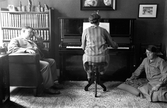 En kvinna sitter vid pianot och spelar. En man i en fåtölj och en kvinna vid en kudde  på golvet sitter och lyssnar.