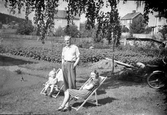 En man står mellan en kvinna och ett barn som sitter i solstolar. Några bostadshus och ett potatisland syns i bakgrunden.