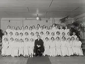 Prästen Aronius (sittande i mitten) med konfirmander, endast flickor. Till höger bakom flickorna syns en stor palmliknande krukväxt. Från vänster sittande: Greta Antonsson, Ingrid Paulsson, Inga Olausson, Lilly Bondesson, May Johansson, Marianne Nihlen, Maj Berntsson, Maj Eliasson, Mildred Lövgren, Signe Carlsson, Laga Hedberg, Ingrid Carlsson, Birgit Johansson 2:a raden: Ingrid Gunnarsson, Birgit Strandberg, Inga-Lisa Andersson, Anna-Lisa Andersson, Rut Johansson, Ingegärd Johansson, Lillan Johansson, Alice Carlsson, Elin Bengtsson, Maud Martinsson, Gunnel Carlsson, Anna Svensson, Herdis Andersson 3:e raden: Evy Granat, Karin Dahlstrand, Karin Rydberg, Kerstin Svensson, Hulda Johansson, Solveig Johansson, Elsa Sandberg, Kerstin Olsson, Inga Gustavsson, Sonja Andersson, Rut Svensson, Anna-Stina Johansson, Dagmar Thoresson, Thordis Johansson 4:e raden: Anna-Lisa Carlsson, Ingeborg Albrektsson, Anna-Greta Larsson, Gulli Antonsson, Kerstin Backman.