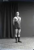 Porträtt, man stående i helfigur iklädd brottarkläder, med många medaljer på bröstet. Ateljébild.
