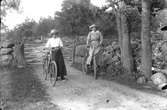Jenny och Berta Andersson, Drängsered, med varsin cykel framför en gärdsgård. En av dem cyklar och den andra leder sin cykel. Båda kvinnorna  är klädda i lång kjol och hatt.