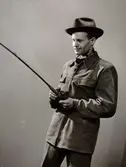 Manlig modell iklädd hatt och moccajacka från C. O. Ericson, Malung, håller i fiskespö. Lädermarknaden, Nordiska Kompaniet 1949, 