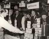 Försäljning av italienska produkter. Kunder framför försäljningsbord, manlig expedit i vit skjorta och förkläde. 