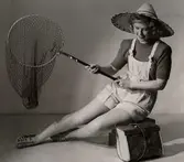 Sommarmode från varuhuset Nordiska Kompaniet i Stockholm 1944. En kvinnlig modell iklädd korta hängselbyxor, tröja, stråhatt och sandaler sitter med en fiskehåv i handen. Vid hennes sida står en väska av flätat näver.