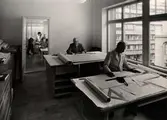 Inredningsavdelningens ritkontor på varuhuset Nordiska Kompaniet i Stockholm 1947. Två män sitter vid varsitt skrivbord och arbetar med ritningar. Ljuset faller in genom ett stort fönster. Bakom dem ser man genom en dörröppning in i ytterligare ett kontorsrum där skjortklädda män arbetar.