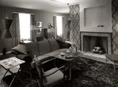Interiör med vardagsrumsmöbler från varuhuset Nordiska Kompaniet i Stockholm 1947. Framför den öppna spisen en soffa, fåtölj och dukat kaffebord samt ett brickbord med keramiköverdel. på golvet en mönstrad ryamatta. Stormönstrade tapeter med bladrankor.