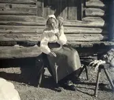 Spånad på slända, Orsa. En kvinna sitter utanför en liggtimrad byggnad och spinner ullgarn på hängande slända.