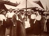 Marknadsdag i Fagersta. En grupp kvinnor i tidstypisk klädsel rör sig kring marknadsstånden.