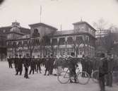 Alhambra, restaurang och teater, på Djurgårdsslätten, Djurgården, Stockholm. Uppfört av hovkonditor Pohl omkring 1830. Även kallat Pohlsro. Byggnadens fasad i bakgrunden. En folksamling - män, barn, kvinnor, cyklar mm står framför på grusplanen. 