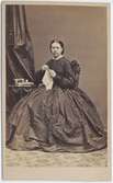 Kabinettkort. Porträtt, helfigur, av kvinna. Hon är iklädd mörk klänning med vid kjol, vida ärmar och liten vit krage. Hon sitter vid ett litet pelarbord och håller ett broderi i händerna. Dräktbilder dam.  1860-tal.