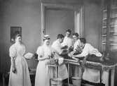 Behandling pågår, en patient ligger på ett operationsbord medan några läkare tar hand om honom. I förgrunden står två sjuksköterskor. Interiör från Serafimerlasarettet 1898.