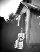 Porträtt av en kvinna i vit blus med fårbogsärmar, mörk kjol och randigt förkläde som sitter på en trappa.