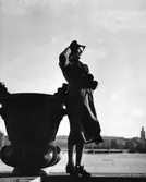 Modell klädd i öppen kappa, snörskor, handskar, och hatt. Bär handväska under armen. I högra hörnet skymtar en person klädd i hatt med cigarett i handen. Miljö: stadshuskajen i Stockholm.