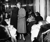 Modevisning i lunchrummet på Nordiska Kompaniet. Modell klädd i regnkappa, hatt och klackskor med öppen tå visar upp kappans insida för åskådarna.