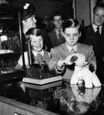 Pianisten Pierino Gamba på besök på Nordiska Kompaniet. Gamba tittar på en leksakshund som står på disken. I bakgrunden står två pojkar, en man och en kvinna. Kvinnan bär hatt med flor.