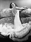 Lilian Harvey - utställning på Nordiska Kompaniet. Modell klädd i vit balklänning med volanger och blomdekorationer. Står vid en blommig soffa.