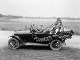 Brudpar i bil dekorerad med löv och blommor. Chauffören finns också med på bilden.