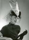 Porträtt av kvinna klädd i hatt med flor och vita fjädrar, från Margit Roth, samt mörk överdel och mörka handskar.