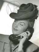 Porträtt av kvinna klädd i hatt med dekorband och flor, kavaj och treradigt pärlhalsband, håller i en telefonlur.